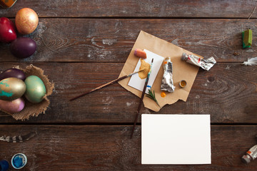 Blank greeting card with colorful eggs flat lay. Top view on wooden table with empty white paper and art tools for decorating eggs for Easter, free space for text