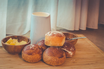 Breakfast with freshly baked bread buns covered in flour, butter and milk on wooden board by kitchen window curtain.