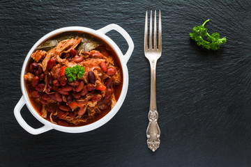 Beans with pork stewed in spicy tomato sauce with onion, paprika, beer, bell and pink pepper. White soup bowl, vintage fork and fresh parsley on black stone, top view.