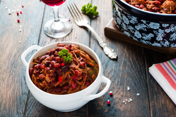Beans with pork stewed in spicy tomato sauce with onion, paprika, beer, bell and pink pepper. White soup bowl, casserole, glass of red wine and vintage fork on wooden rustic table.