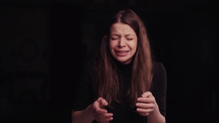 Young European brunette girl with a messy hair in a black turtleneck on the black background. Attractive upset woman is loudly crying, covering her face with hands. Arguing, having conflict.