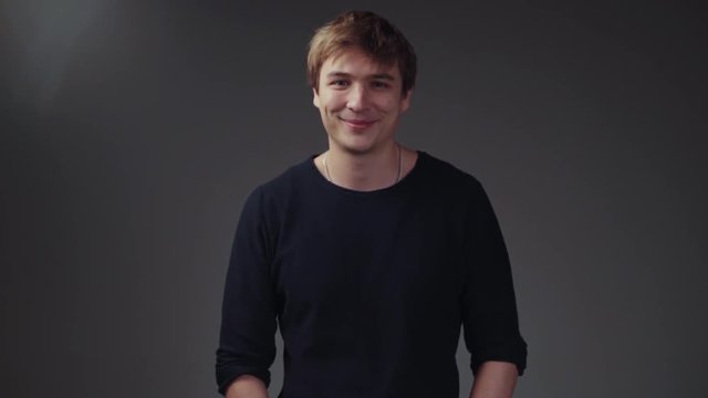 Handsome young European man in a stylish black sweater on the grey screen. Funny boy looking down, lifts his head and looks towards the camera, gives a cute playful smile.