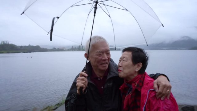 Senior Asian couple under clear umbrella while travel to cloudy lake, explore the world together after retire