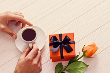 Young man waiting in the cafe with orange present and orange rose. Drinking coffee