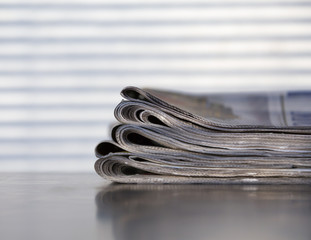 Stack of newspapers on a wooden surface. Curtain shadows in the background.