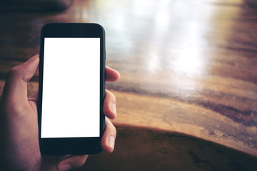 Mockup image of hands holding black mobile phone with blank white screen and vintage wood table background