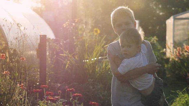 Older Brother Holds Of Young Brother Goes Outdoor Next To The Flowers In The Park During Sunset With Lens Flare Effects In Slowmotion. 1920x1080