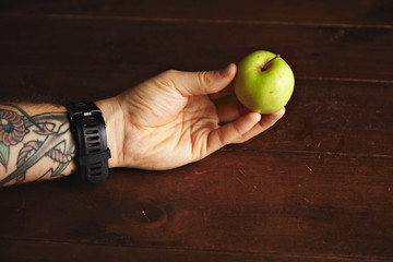 Tattooed man's hand holds small sweet green apple above wooden table
