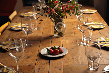 Plate with big fresh strawberries isolated in center of served for holiday dinner table, between empty dishes and wine glasses, in front of big jar with branches of eucalyptus and rowan inside