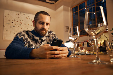 Thoughtful bearded man in warm wool knitted icelandic sweater with horses using his smart phone, sitting in served table for holiday dinner, warm lit ambient lightning inside big spacious room