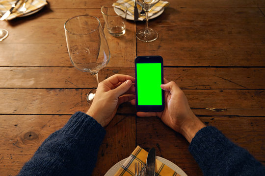 Top view on man's hand using smart phone above served table in restaurant, next to empty wine glass. Screen filled with chromakey green