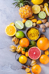 Variety of whole and sliced citrus fruits pineapple, grapefruit, lemon, lime, kumquat, clementine and physalis with mint and yellow spices and nuts over gray textured background. Top view with space. 