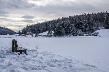 Fototapeta premium A wooden bench at the edge of a lake covered entirely by the snow. In the background, a mountain with trees. Cloudy weather