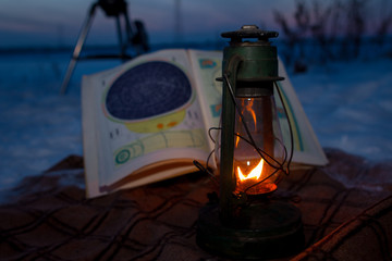 Fototapeta na wymiar Burning oil lamp standing on a map of the sky in the winter on a background of sunset sky and telescope