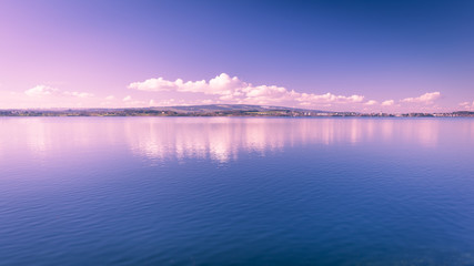 Fototapeta premium Clear day. Lake Zug in central Switzerland. Clouds are reflected in water lake. At the edge water is clear and see the bottom of the lake.