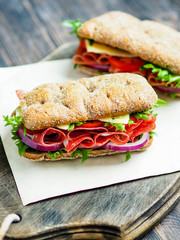 Two fresh sandwiches with jamon, cheese, tomato, lettuce and onion in whole-grain biscuits on a dark wood board on a dark background

