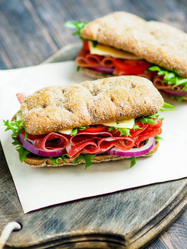 Two fresh sandwiches with jamon, cheese, tomato, lettuce and onion in whole-grain biscuits on a dark wood board on a dark background

