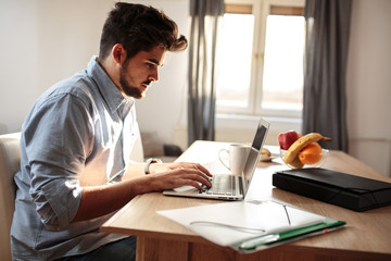 Young man sitting at the morning in his living room and using laptop.Watching online stock market.