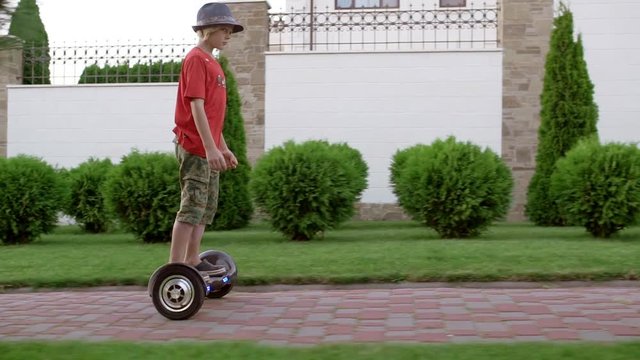 Young Caucasian boy in red t-shirt military shorts and hat driving gyroscooter through alley in slowmotion