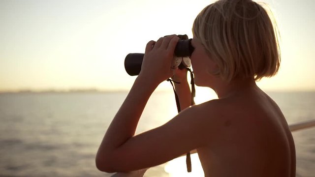 Topless Caucasian blonde boy looking at sea through binoculars in slowmotion