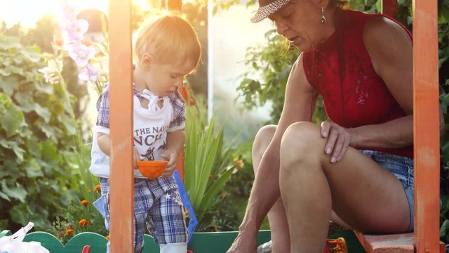Fashionable grandma in hat and grandson playing in the sandbox, molded figurines from sand at sunset time through the sun with lense flare effects. Happy childhood. 3840x2160