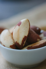 Heap of brazil nuts in white ceramic bowl over the brown fabric.