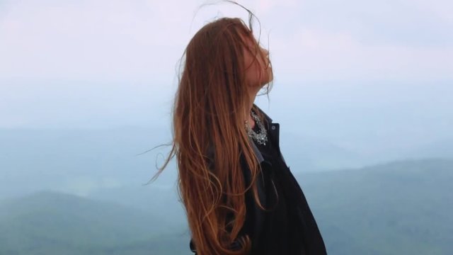 Amazing young lady with long hair in a black stylish dress, and gorgeous leather jacket. Beautiful European girl standing on the top of the hill, strong wind is playing with her hair.