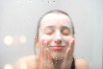 Fototapeta na wymiar Close-up portrait of a woman with soap on her wet face standing behind the glass in the shower. Image with soft focus