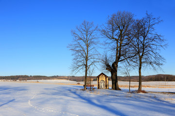 Kapliczka z ławeczką na pustkowiu w zimowym krajobrazie. - obrazy, fototapety, plakaty
