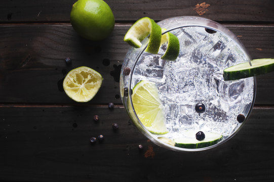 Glass of gin tonic with ice, cucumber, lime over a dark wood table