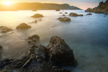 Long exposure seascape at sunrise. View of the cliff into the sea and distant islands. In the backlight sunbeam light. Paleokastrica. Corfu. Ionian archipelago. Greece.