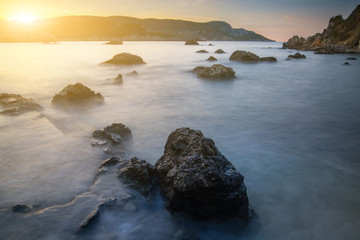 Fototapeta na wymiar Long exposure seascape at sunrise. View of the cliff into the sea and distant islands. In the backlight sunbeam light. Paleokastrica. Corfu. Ionian archipelago. Greece.