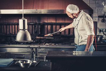 Young white chef in blue apron and hat standing near the brazier whith coals. Man cooking beef steak in the interior of modern professional kitchen