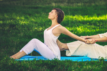 Young man and woman dressed in a white robe doing thai massage with yoga exercises sitting on the mat. Sunny summer park with green lawn in the background