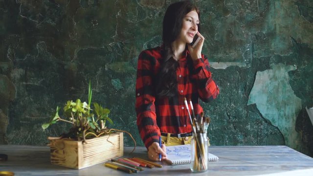 Young woman artist painting scetch on paper notebook with pencil. Girl smile and talks phone