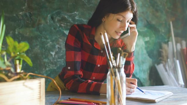 Young woman artist painting scetch on paper notebook with pencil. Girl smile and talks phone