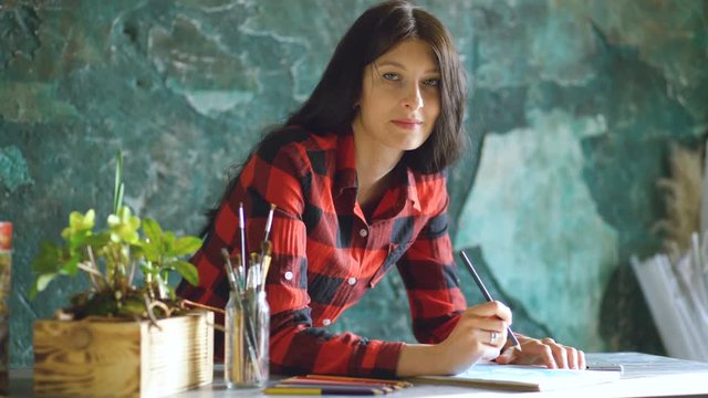Young woman artist painting scetch on paper notebook with pencil and looks into camera smiling