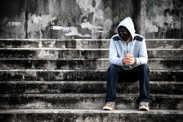 Asian man in the white hood and black mask with confused depression sitting on stairs, in scary abandoned building, The concept of unemployed, sadness, depressed and human problems in dark tone.