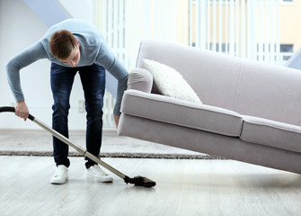 Young man with vacuum cleaner cleaning floor at home - obrazy, fototapety, plakaty