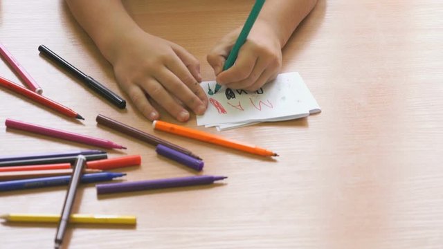 Kid sitting at the desk draws the image on the paper sheet using the colored felt pens. Close-up