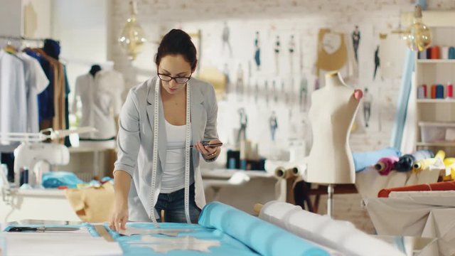 Beautiful Brunette Fashion Designer Lays Out Templates on the fabrics and Consults Her Smartphone. She Works in a Light Colorful Studio Full of Various Clothes, Fabrics and Sketches on the Wall.