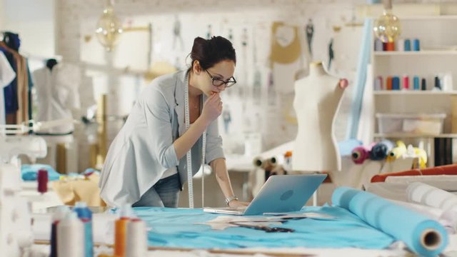  Beautiful Female Designer Uses Laptop on Her Desk that is Covered with Various Fabrics and Sewing Items. Her Studio is Sunny, There is Mannequin, Clothes Hanging and Sketches Pinned to the Wall. 