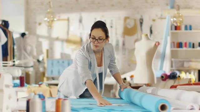 Beautiful Female Fashion Designer Straightens Roll of Blue Fabrics and Lays Out Templates on it.  She Works in a Light Colorful Studio Full of Various Clothes, Fabrics and Sketches on the Wall. 