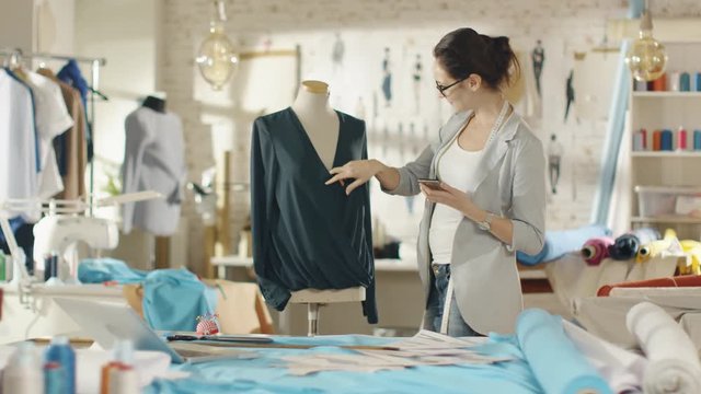 Beautiful Woman Fashion Designer Adjusts Blouse on a Mannequin, Uses Her Smartphone and Smiles. Her Studio is Bright and Sunny, Clothes Hanging, Colourful Fabrics Lying on the Table. 
