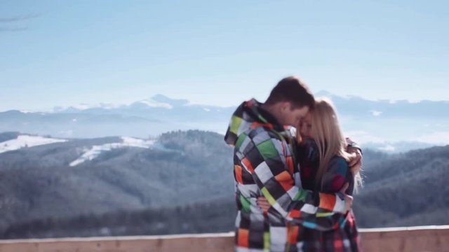 Young man tenderly embracing his attractive blonde girlfriend while standing on the mountain observation deck. Amazing scenery on the background. Romantic atmosphere. True love.