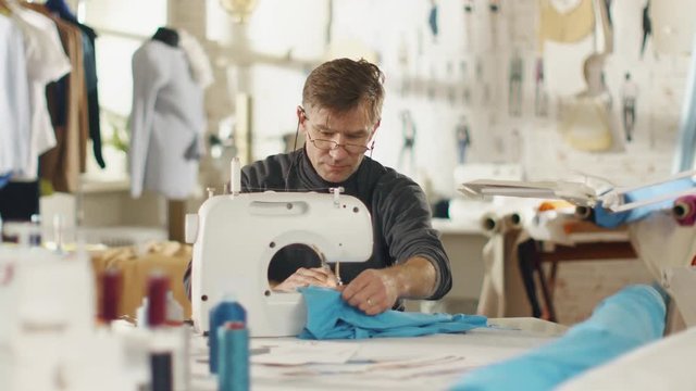 Male Fashion Designer/ Dressmaker Working on a Sewing Machine in Him Sunny Studio. Various Sewing items and Fabrics Laying Around Him, Visible Sketches Pinned to the Wall.  Shot on RED EPIC 4K (UHD).