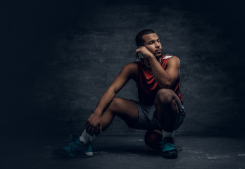 A black basketball player sits on a floor and holds basket ball.
