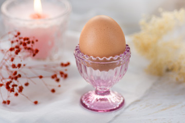Easter egg in pink crystal cup,white linen cloth background,burning candle,bouquet of spring delicate beige and red flowers,decoration,pastel colors,top angle view