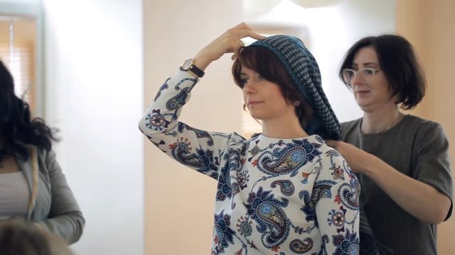 stylist shows on model how to tie scarf on your head. It is group master class on style. We see brunette with kare in jeans, white sweater with fancy pattern. Behind her there is consultant adult