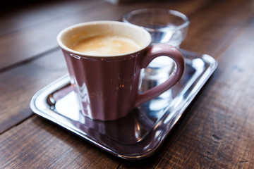 A pink cup of black coffee and a glass of water on a silver tray. Selective focus, small depth of field.